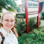 Leah, the blog author, a 20-something blond woman wearing a pink hair scarf smiles in front of a museum sign reading "Safe Haven"